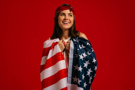 Téléchargez les photos : Jolie jeune femme hipster portant le drapeau américain sur les épaules et souriant sur fond rouge - en image libre de droit