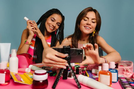 Foto de Dos jóvenes vloggers de belleza se preparan para transmitir en vivo mientras se sientan en el escritorio contra el fondo azul - Imagen libre de derechos