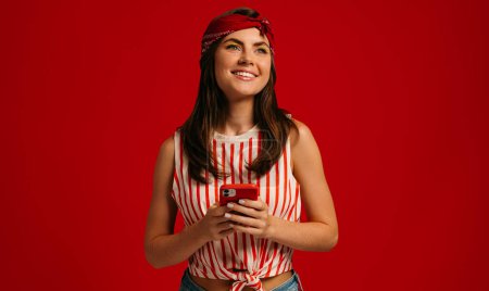 Téléchargez les photos : Élégant jeune femme hipster tenant téléphone intelligent et souriant tout en se tenant debout sur fond rouge - en image libre de droit