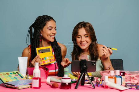 Foto de Dos animadas presentadoras femeninas transmitiendo vlog de belleza mientras están sentadas en el escritorio contra el fondo azul - Imagen libre de derechos
