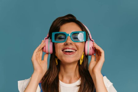 Téléchargez les photos : Portrait de jeune femme joyeuse dans des écouteurs et des lunettes à la mode profitant de la musique sur fond bleu - en image libre de droit
