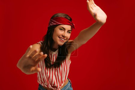 Téléchargez les photos : Joyeux jeune femme hipster étirant les mains et souriant tout en se tenant debout sur fond rouge - en image libre de droit