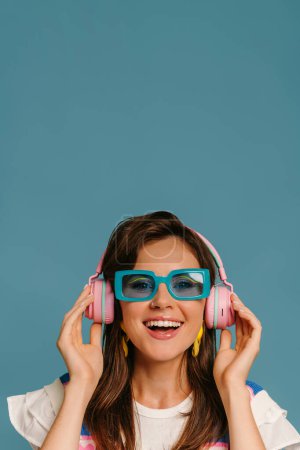 Téléchargez les photos : Portrait de jeune femme heureuse dans des écouteurs et des lunettes à la mode profitant de la musique sur fond bleu - en image libre de droit