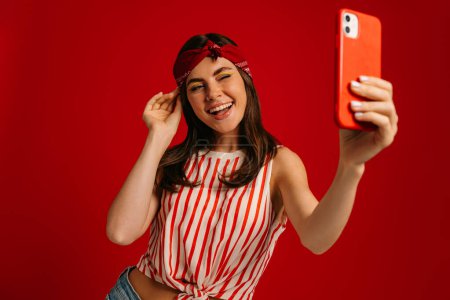 Téléchargez les photos : Joyeux jeune femme hipster faire selfie par téléphone mart et clin d'oeil tout en se tenant debout sur fond rouge - en image libre de droit