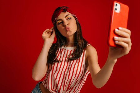 Foto de Juguetona joven hipster mujer haciendo selfie por teléfono del mercado, mientras que de pie sobre fondo rojo - Imagen libre de derechos