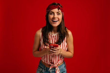 Téléchargez les photos : Joyeuse jeune femme hipster tenant téléphone intelligent et souriant tout en se tenant debout sur fond rouge - en image libre de droit