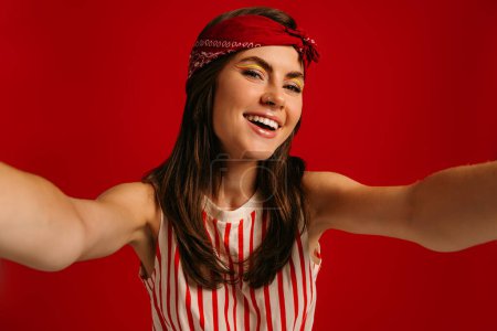 Téléchargez les photos : Joyeux jeune femme hipster faire selfie tout en gardant le bras tendu sur fond rouge - en image libre de droit