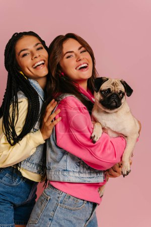 Téléchargez les photos : Deux belles jeunes femmes collant et souriant tout en portant chien carlin mignon sur fond rose - en image libre de droit