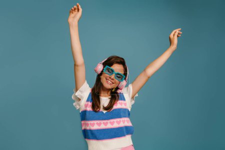 Téléchargez les photos : Joyeux jeune femme dans des écouteurs et des lunettes à la mode profitant de la musique tout en dansant sur fond bleu - en image libre de droit