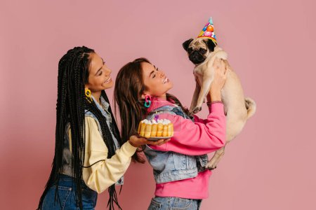Téléchargez les photos : Deux jeunes femmes heureuses collant tout en portant chien chiot mignon et gâteau d'anniversaire sur fond rose - en image libre de droit