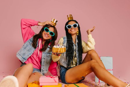 Téléchargez les photos : Deux jeunes femmes ludiques tenant gâteau d'anniversaire et souriant tout en étant assis près de boîtes-cadeaux sur fond rose - en image libre de droit