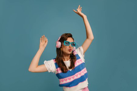 Téléchargez les photos : Belle jeune femme dans des écouteurs et des lunettes à la mode profitant de la musique tout en dansant sur fond bleu - en image libre de droit