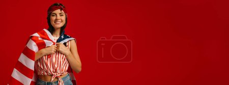 Téléchargez les photos : Joyeux jeune femme hipster portant drapeau américain sur les épaules tout en se tenant sur fond rouge - en image libre de droit