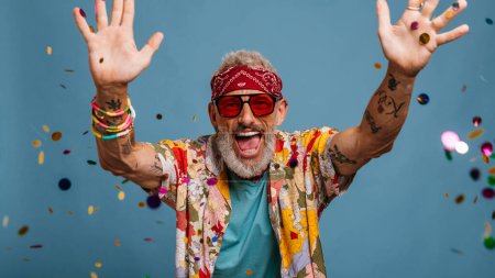 Foto de Hombre mayor alegre en camisa funky y bandana lanzando confeti colorido sobre fondo azul - Imagen libre de derechos