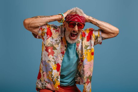 Téléchargez les photos : Choqué hipster senior homme en élégant chemise funky tenant la tête dans les mains et criant sur fond bleu - en image libre de droit