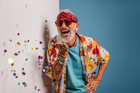 Photo for Happy senior man in funky shirt and bandana blowing colorful confetti on white and blue background - Royalty Free Image