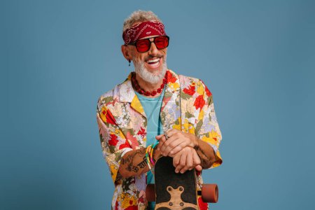 Téléchargez les photos : Heureux homme mature en chemise funky appuyé sur longboard et souriant sur fond bleu - en image libre de droit