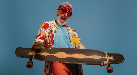 Photo for Playful senior man carrying longboard like a guitar and feeling like a rock star against blue background - Royalty Free Image