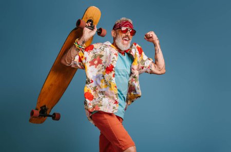 Cool senior man in funky shirt carrying longboard and smiling against blue background