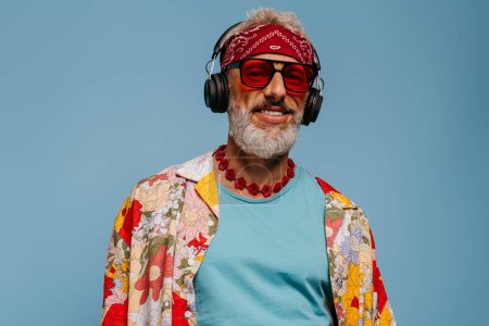 Téléchargez les photos : Homme âgé confiant en chemise funky et écouteurs écoutant la musique sur fond bleu - en image libre de droit