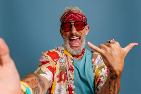 Foto de Hombre mayor fresco en camisa funky con estilo y gafas de moda haciendo selfie y gestos sobre fondo azul - Imagen libre de derechos