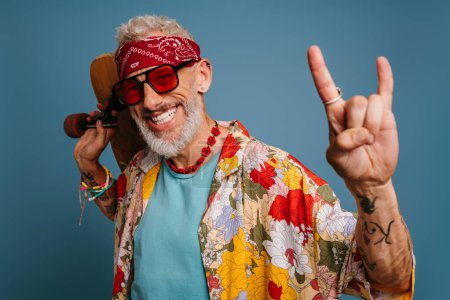 Photo for Hipster mature man in stylish funky shirt carrying longboard and gesturing against blue background - Royalty Free Image