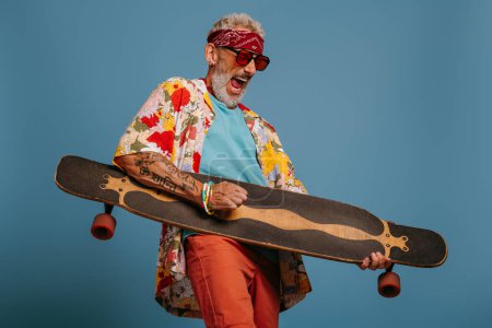 Foto de Hombre mayor juguetón llevando longboard como una guitarra y sintiéndose como una estrella de rock sobre fondo azul - Imagen libre de derechos