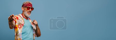Foto de Feliz hombre mayor hipster en elegante camisa funky apuntando cámara contra fondo azul - Imagen libre de derechos
