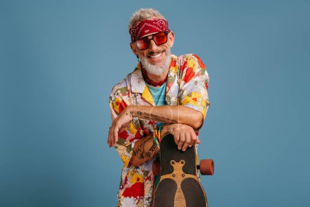 Téléchargez les photos : Hipster homme mature en chemise funky élégant appuyé sur longboard et souriant sur fond bleu - en image libre de droit