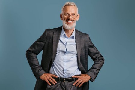 Foto de Confiado hombre de negocios maduro cogido de la mano en la cadera y sonriendo mientras está de pie sobre fondo de color - Imagen libre de derechos