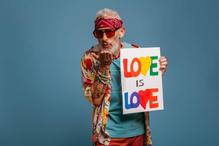 Foto de Alegre hipster senior hombre en funky camisa sosteniendo pancarta y soplando un beso sobre fondo azul - Imagen libre de derechos
