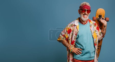 Téléchargez les photos : Cool senior homme en chemise funky portant longboard et souriant sur fond bleu - en image libre de droit