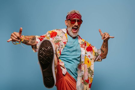 Téléchargez les photos : Cool hipster senior homme en chemise funky élégant coup de pied jambe et geste sur fond bleu - en image libre de droit