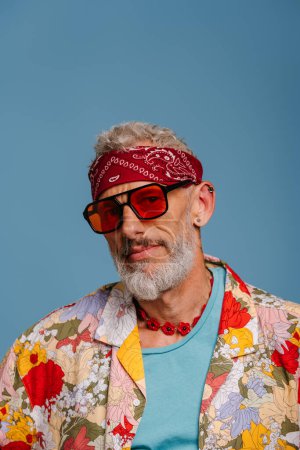 Téléchargez les photos : Portrait de hipster senior homme en chemise funky élégant et des lunettes à la mode regardant la caméra sur fond bleu - en image libre de droit