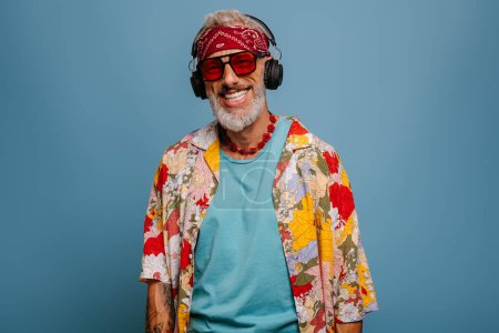 Foto de Hombre mayor alegre en camisa funky y auriculares escuchando la música contra el fondo azul - Imagen libre de derechos
