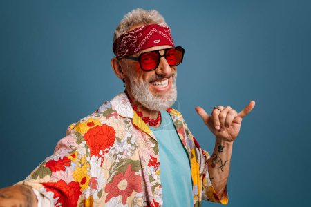 Foto de Hipster hombre senior en elegante camisa funky y gafas de moda haciendo selfie y gestos sobre fondo azul - Imagen libre de derechos
