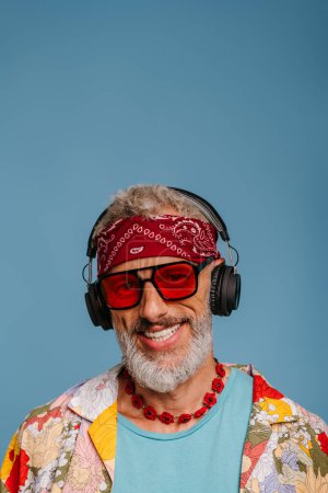 Foto de Hombre mayor alegre en camisa funky y auriculares escuchando la música sobre fondo azul - Imagen libre de derechos