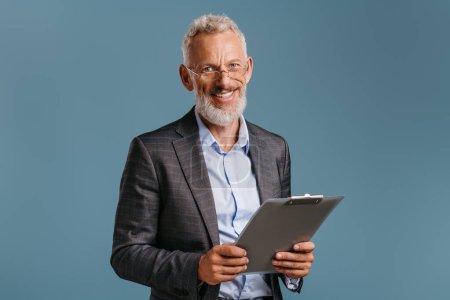 Téléchargez les photos : Homme d'affaires mature confiant tenant dossier de fichier et souriant tout en se tenant debout sur fond coloré - en image libre de droit