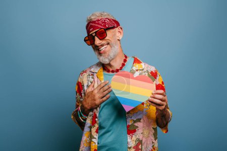 Téléchargez les photos : Joyeux hipster senior homme en chemise funky tenant drapeau arc-en-ciel en forme de coeur sur fond bleu - en image libre de droit