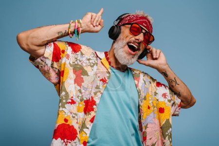 Photo for Joyful senior man in funky shirt and headphones listening to the music and dancing on blue background - Royalty Free Image