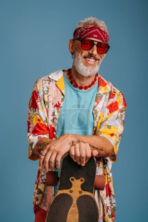 Photo for Hipster senior man in stylish funky shirt leaning on longboard and smiling against blue background - Royalty Free Image