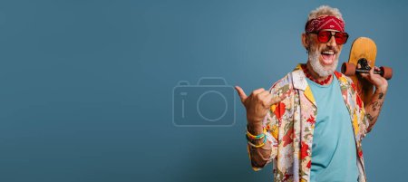Téléchargez les photos : Joyeux hipster senior homme en chemise funky élégant portant longboard et geste sur fond bleu - en image libre de droit