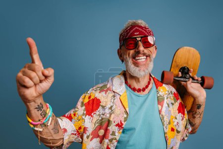 Foto de Alegre hipster hombre maduro en elegante camisa funky llevando longboard y gestos sobre fondo azul - Imagen libre de derechos