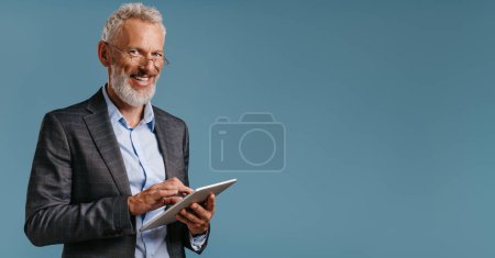 Téléchargez les photos : Homme d'affaires mature confiant travaillant sur tablette numérique et souriant tout en se tenant debout sur fond coloré - en image libre de droit