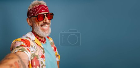 Téléchargez les photos : Hipster senior homme en chemise funky élégant et des lunettes à la mode faisant selfie et souriant sur fond bleu - en image libre de droit