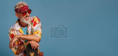 Photo for Stylish mature man in funky shirt leaning on longboard and smiling against blue background - Royalty Free Image