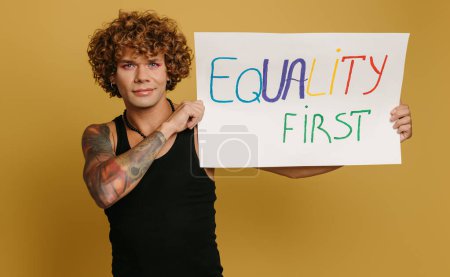 Téléchargez les photos : Beau jeune gay l'homme avec maquillage montrant coloré bannière sur jaune fond - en image libre de droit