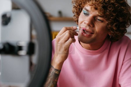 Foto de Confiado joven gay hombre aplicando lápiz labial mientras streaming online maquillaje vlog desde casa - Imagen libre de derechos