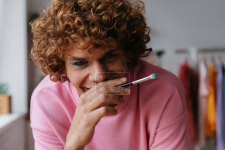 Téléchargez les photos : Timide jeune gay homme tenant maquillage brosse et sourire tout en appliquant fard à paupières à la maison - en image libre de droit