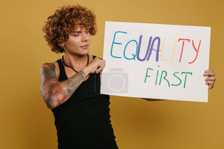 Téléchargez les photos : Beau jeune homme gay avec maquillage tenant bannière colorée sur fond jaune - en image libre de droit